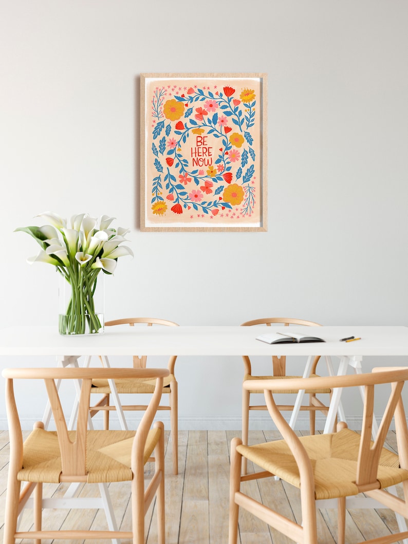 May include: A framed print with a floral design and the text "BE HERE NOW" in red and white lettering. The flowers are in shades of blue, pink, yellow, and orange.