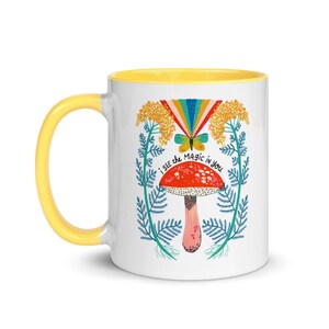 May include: White ceramic mug with a yellow handle and interior. The mug features a whimsical design with a red mushroom, a butterfly, and the text "I see the magic in you". The design is framed by floral elements.