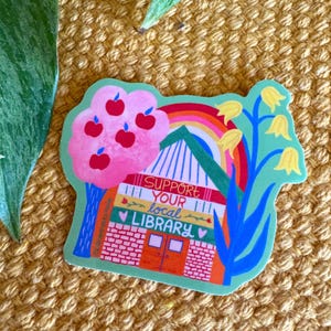May include: A green sticker with a colorful illustration of a library building with a rainbow, apple tree, and flowers. The text "SUPPORT YOUR local LIBRARY" is written on the building.