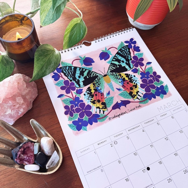 May include: A spiral-bound calendar featuring a colorful illustration of a butterfly with blue, yellow, and red wings, surrounded by purple flowers. The calendar is open to March, with the word "March" and the days of the week visible.