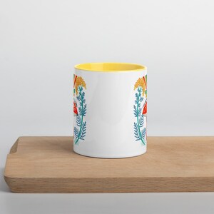May include: White ceramic mug with a yellow interior, featuring a colorful botanical design of red mushrooms, yellow flowers, and blue foliage. The mug is placed on a wooden cutting board.