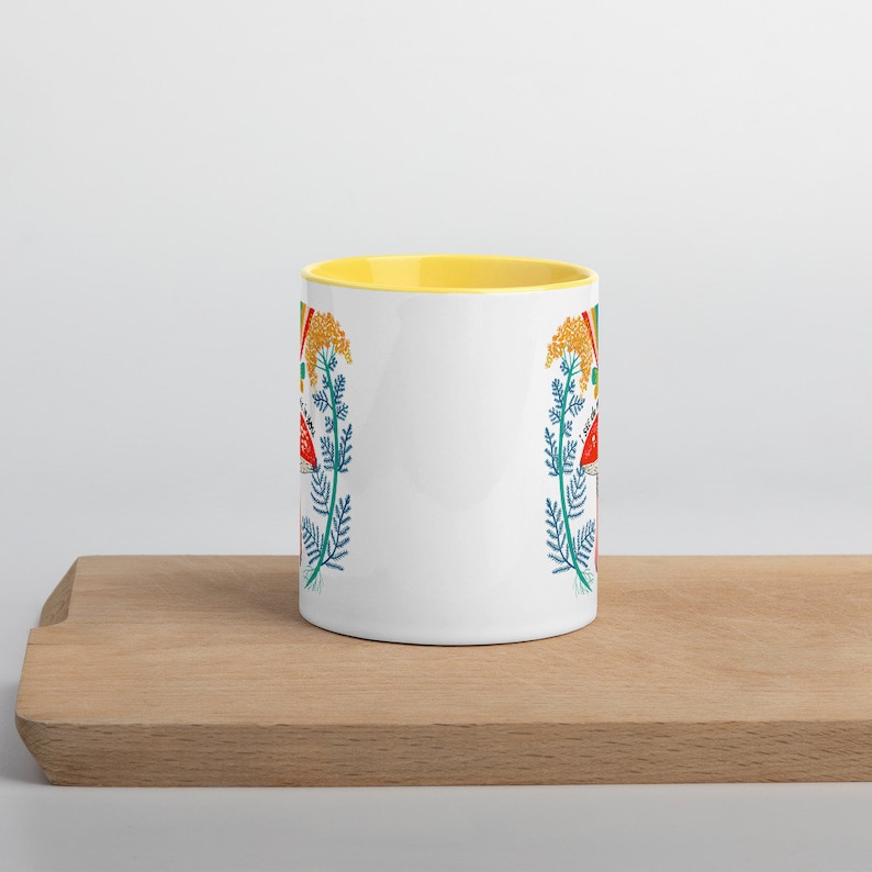 May include: White ceramic mug with a yellow interior, featuring a colorful botanical design of red mushrooms, yellow flowers, and blue foliage. The mug is placed on a wooden cutting board.