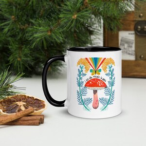 May include: White ceramic mug with a black handle and interior. The mug features a colorful illustration of a red mushroom with white spots, a butterfly, and the text "I see the magic in you."