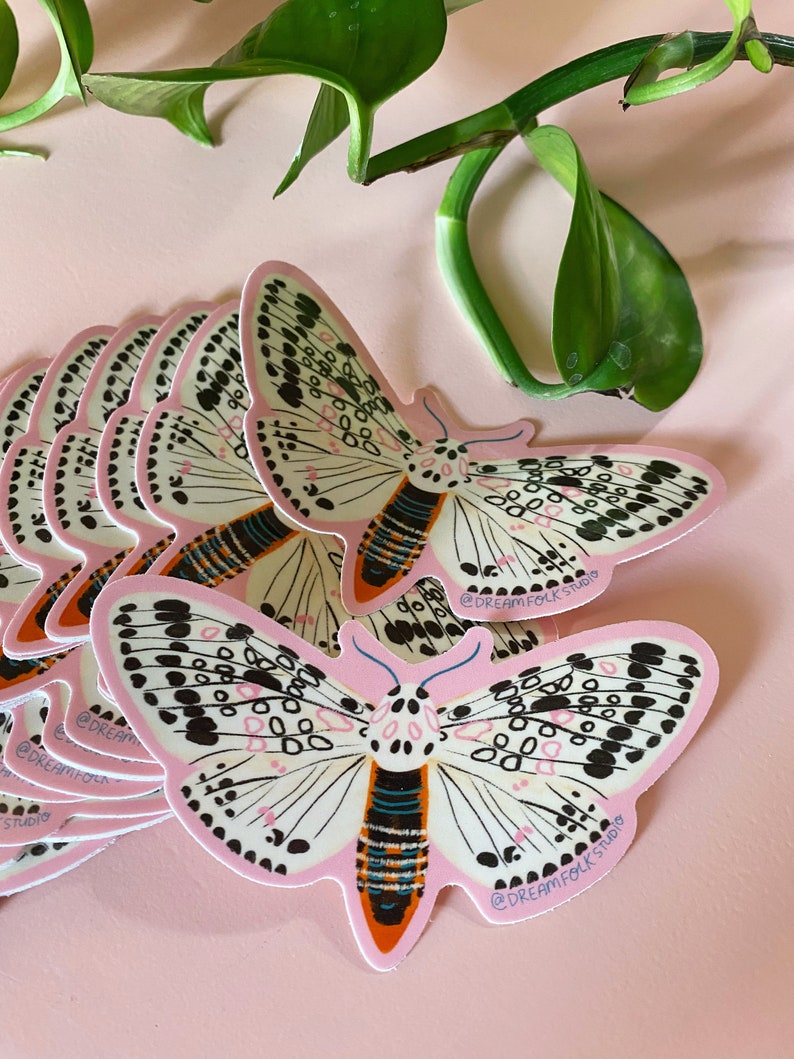 May include: A collection of pink stickers featuring a moth design. The moth has white wings with black spots and a pink outline. The body is orange and black. The stickers are on a pink surface with green leaves in the background. The text "@DREAMFOLKSTUDIO" is on the stickers.