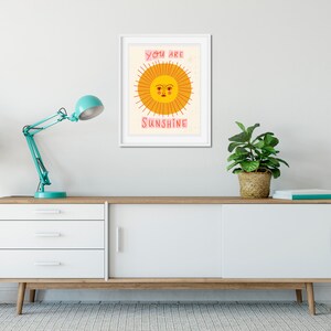 K&ouml;nnte beinhalten: Ein wei&szlig;er gerahmter Druck mit einer gelben Sonne mit einem rosafarbenen Gesicht und dem Text "You are Sunshine" in Rosa.