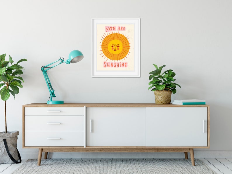 K&ouml;nnte beinhalten: Ein wei&szlig;er gerahmter Druck mit einer gelben Sonne mit einem rosafarbenen Gesicht und dem Text "You are Sunshine" in Rosa.