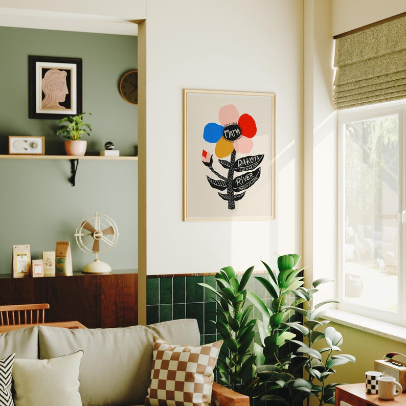 May include: A framed art print featuring a stylized flower design with colorful petals in pink, red, blue, and yellow. The black stem has the words "Mama", "Dakota", and "River". The print is displayed in a bright, well-lit room with plants.