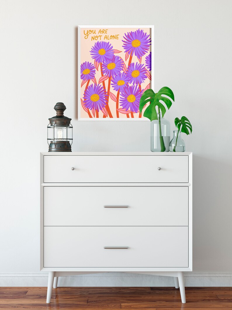 May include: A white framed print with a pink background and purple flowers. The text "You are not alone" is written in orange on the print.