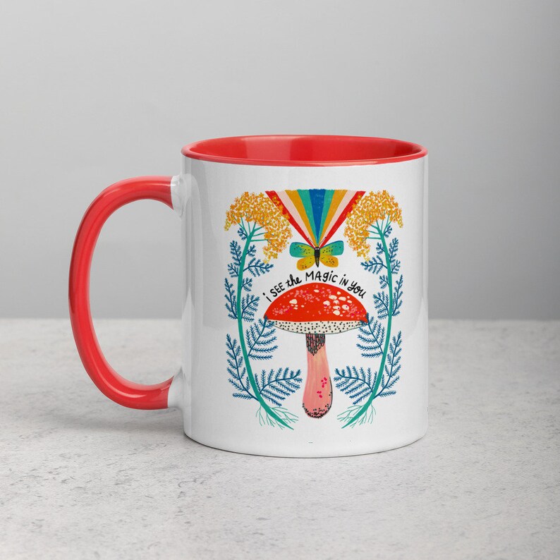 May include: White ceramic mug with a red interior and handle. The mug features a colorful illustration of a mushroom, butterfly, and rainbow, with the text "I SEE THE MAGIC IN YOU". The design is surrounded by floral elements.