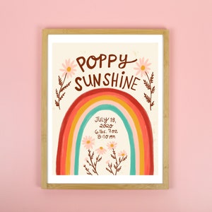 May include: A framed art print with the words "Poppy Sunshine" above a rainbow illustration. The rainbow is in shades of red, orange, yellow, and blue. Below the rainbow are the words "July 18, 2020, 6 lbs. 7 oz, 8:10 am". The print is in a light wood frame.