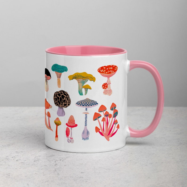 May include: A white ceramic mug with a pink rim and handle. The mug features a colourful illustration of various mushrooms, including red, orange, yellow, green, and purple mushrooms.