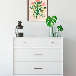May include: A white dresser with three drawers and a framed print of a pink rose bush with green leaves and roots. The dresser is on a wooden floor and there are two green plants in glass vases on top of the dresser.