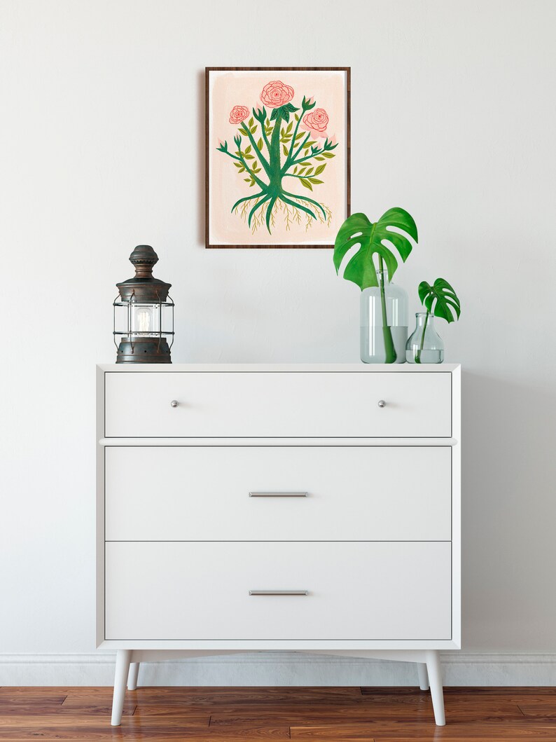 May include: A white dresser with three drawers and a framed print of a pink rose bush with green leaves and roots. The dresser is on a wooden floor and there are two green plants in glass vases on top of the dresser.