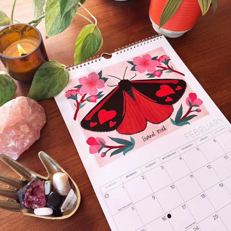 May include: A spiral-bound calendar open to February, featuring a red and black moth illustration with heart-shaped markings. The calendar is surrounded by a lit candle, crystals, and a small dish of stones, suggesting a spiritual or decorative theme.