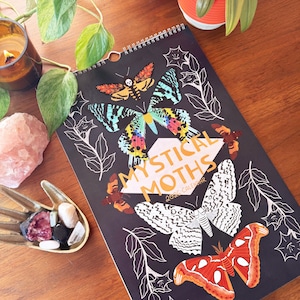 May include: A spiral-bound calendar with the words "Mystical Moths" and "2026 Calendar" on the cover. The cover features colorful moth illustrations on a black background. The moths are in shades of orange, blue, yellow, and white. The calendar is on a wooden surface.