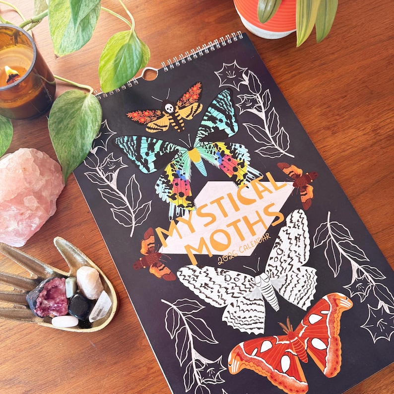 May include: A spiral-bound calendar with the words "Mystical Moths" and "2026 Calendar" on the cover. The cover features colorful moth illustrations on a black background. The moths are in shades of orange, blue, yellow, and white. The calendar is on a wooden surface.
