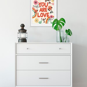 Pu&ograve; includere: Una stampa incorniciata bianca con un design floreale colorato e il testo "YOU ARE LOVE" in lettere rosse.