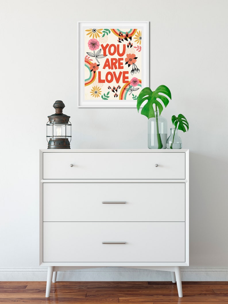 Pu&ograve; includere: Una stampa incorniciata bianca con un design floreale colorato e il testo "YOU ARE LOVE" in lettere rosse.