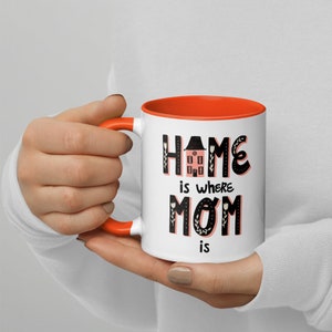 K&ouml;nnte beinhalten: Wei&szlig;e Keramiktasse mit orangefarbenem Rand. Die Tasse zeigt einen schwarzen Text mit der Aufschrift "Home is where Mom is" mit einer rosa Haus-Illustration in der Mitte des Wortes "Home".