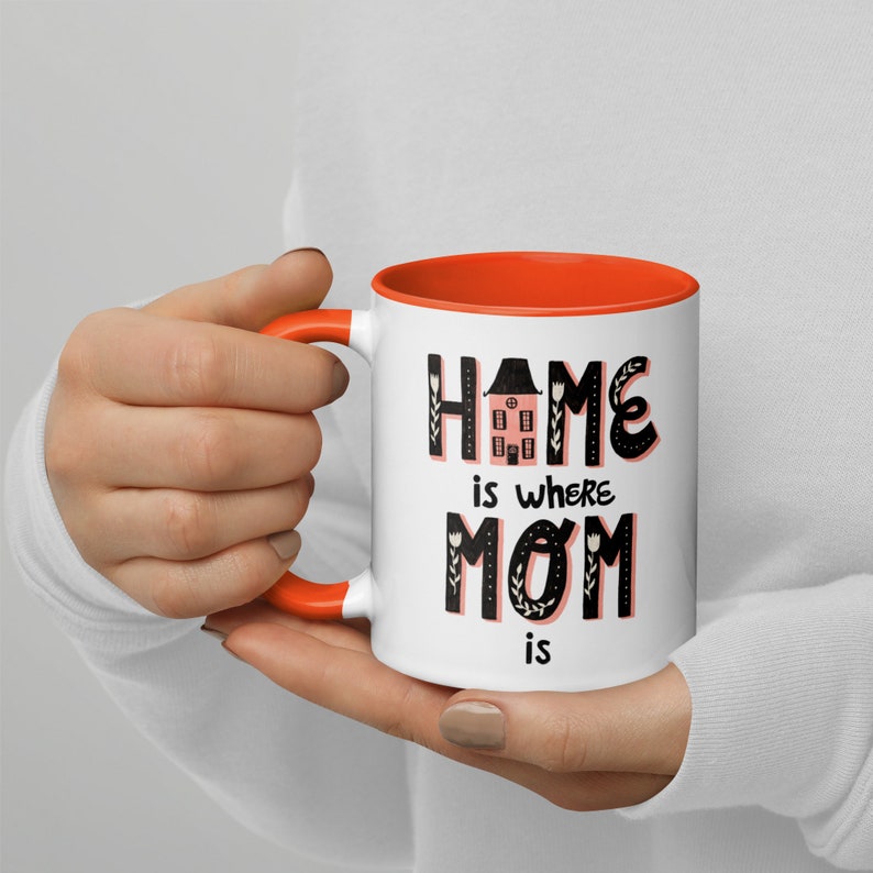 K&ouml;nnte beinhalten: Wei&szlig;e Keramiktasse mit orangefarbenem Rand. Die Tasse zeigt einen schwarzen Text mit der Aufschrift "Home is where Mom is" mit einer rosa Haus-Illustration in der Mitte des Wortes "Home".