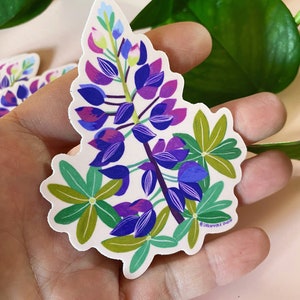 May include: A sticker featuring a purple and blue lupine flower with green leaves. The sticker has a white background and the text "Dreamfolk Studio" in the bottom right corner.