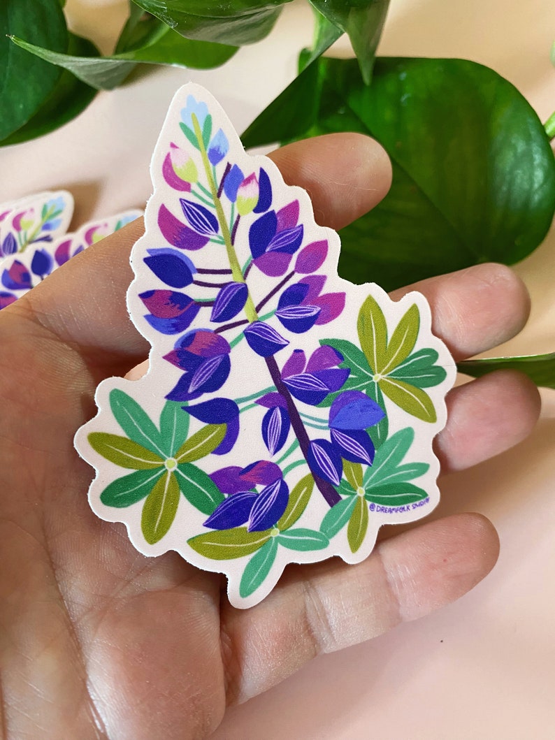 May include: A sticker featuring a purple and blue lupine flower with green leaves. The sticker has a white background and the text "Dreamfolk Studio" in the bottom right corner.