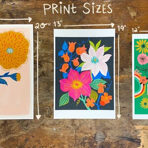 May include: Three different floral prints with white borders. The first print is 16 inches by 20 inches and features a single orange flower with blue leaves on a pink background. The second print is 13 inches by 19 inches and features a cluster of pink, white, and orange flowers on a black background. The third print is 14 inches by 18 inches and features a green background with a rainbow design and the text "You are Love" in orange, red, and yellow letters.