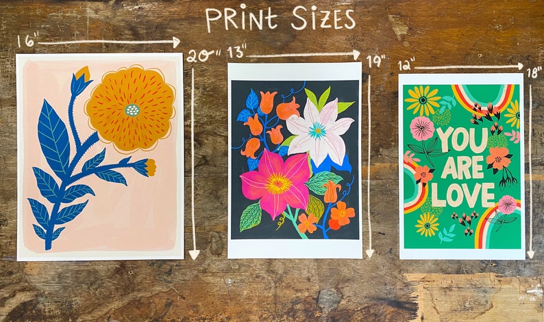 May include: Three different floral prints with white borders. The first print is 16 inches by 20 inches and features a single orange flower with blue leaves on a pink background. The second print is 13 inches by 19 inches and features a cluster of pink, white, and orange flowers on a black background. The third print is 14 inches by 18 inches and features a green background with a rainbow design and the text "You are Love" in orange, red, and yellow letters.