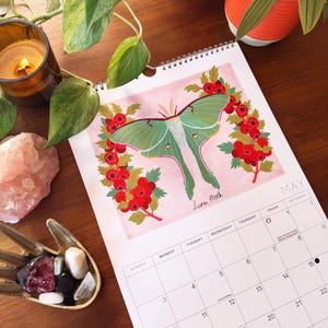 May include: A May calendar page featuring a green Luna moth illustration with red berries and the text "Luna Moth." The calendar is open on a wooden surface, with a lit candle, crystals, and a decorative bowl.