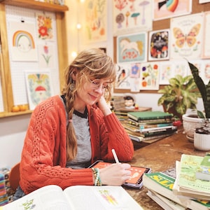 May include: A person wearing a burnt orange cable-knit cardigan over a black and white striped shirt, using a white stylus on a digital tablet. The person has braided hair and wears glasses, with stacks of books and botanical illustrations nearby.