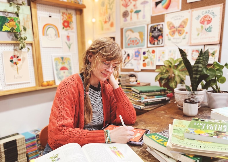 May include: A person wearing a burnt orange cable-knit cardigan over a black and white striped shirt, using a white stylus on a digital tablet. The person has braided hair and wears glasses, with stacks of books and botanical illustrations nearby.