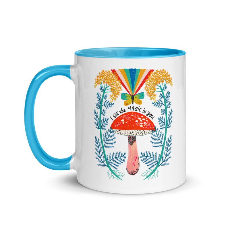 May include: White ceramic mug with a blue handle and interior. The mug features a colorful illustration of a red mushroom with white spots, a rainbow, and the text "I see the magic in you."