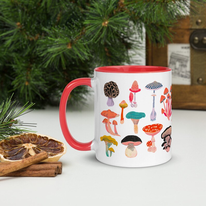 May include: White ceramic mug with a red rim, featuring a colourful illustration of various mushrooms. The mug is filled with a white liquid.