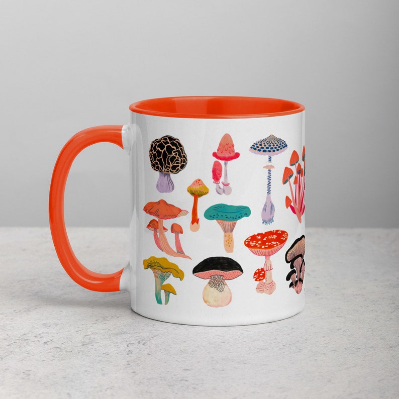 May include: White ceramic mug with an orange rim featuring a colourful illustration of various mushrooms. The mug is on a light grey surface.