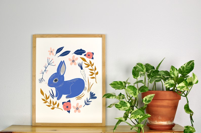 May include: A framed print of a blue rabbit surrounded by a wreath of flowers and leaves. The rabbit is sitting on its hind legs and has a carrot behind it. The flowers are pink, white, and orange.