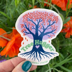 May include: A sticker featuring a stylized tree with red leaves and blue roots. The text "Tree Hugger" is written around the tree in green letters with a purple heart in the center. The sticker is on a white background and has a purple border.