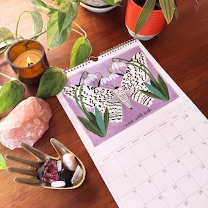 May include: A spiral-bound calendar open to January, featuring a watercolor illustration of a white witch moth with a floral design. The calendar is on a wooden surface, with a lit candle, crystals, and plants.