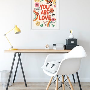 Pu&ograve; includere: Una stampa incorniciata bianca con un design arcobaleno colorato e fiori. Il testo "YOU ARE LOVE" &egrave; in lettere rosse e grassetto.