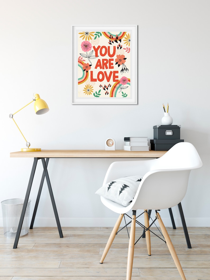 Pu&ograve; includere: Una stampa incorniciata bianca con un design arcobaleno colorato e fiori. Il testo "YOU ARE LOVE" &egrave; in lettere rosse e grassetto.