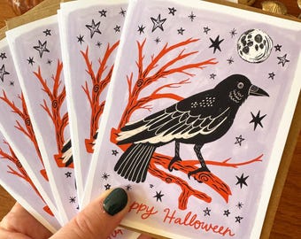 May include: A stack of rectangular greeting cards featuring a black crow perched on a bright orange tree branch against a light purple background. The design includes a white moon, black stars, and the red handwritten text Happy Halloween.