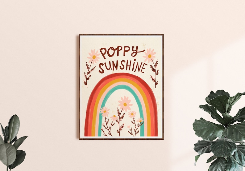 May include: A framed art print featuring a colorful rainbow with the words "Poppy Sunshine" in brown script. The artwork includes daisies and floral accents, set against a cream background. The print is displayed on a light beige wall.