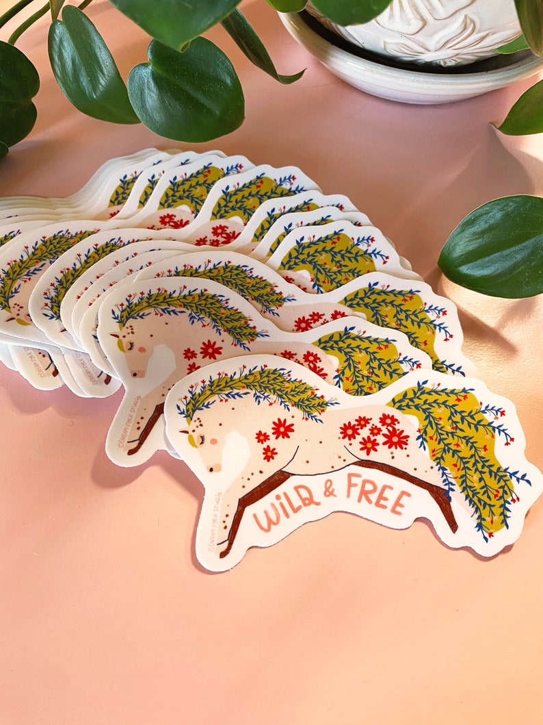 May include: A stack of white stickers with a colorful illustration of a horse with a floral wreath and the text "WILD & FREE".
