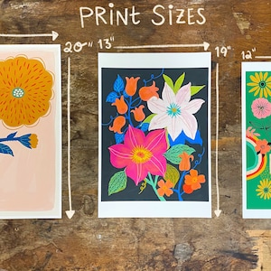 May include: Three different floral prints with white borders. The first print is 16 inches by 13 inches and features a single orange flower with blue leaves on a pink background. The second print is 20 inches by 13 inches and features a cluster of pink, white, and orange flowers on a black background. The third print is 19 inches by 12 inches and features a green background with a rainbow design and the text "You are Love" in orange and white letters.