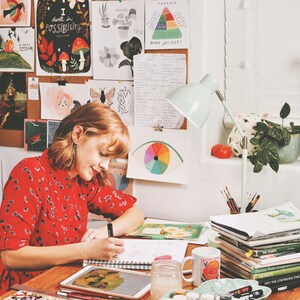 May include: A woman in a red floral dress sits at a desk, sketching in a sketchbook. She has short, brown hair and is wearing a silver earring. The desk is covered in art supplies, including paint, brushes, and a tablet. There are several framed illustrations on the wall behind her, including one that says "I dwell in possibility".
