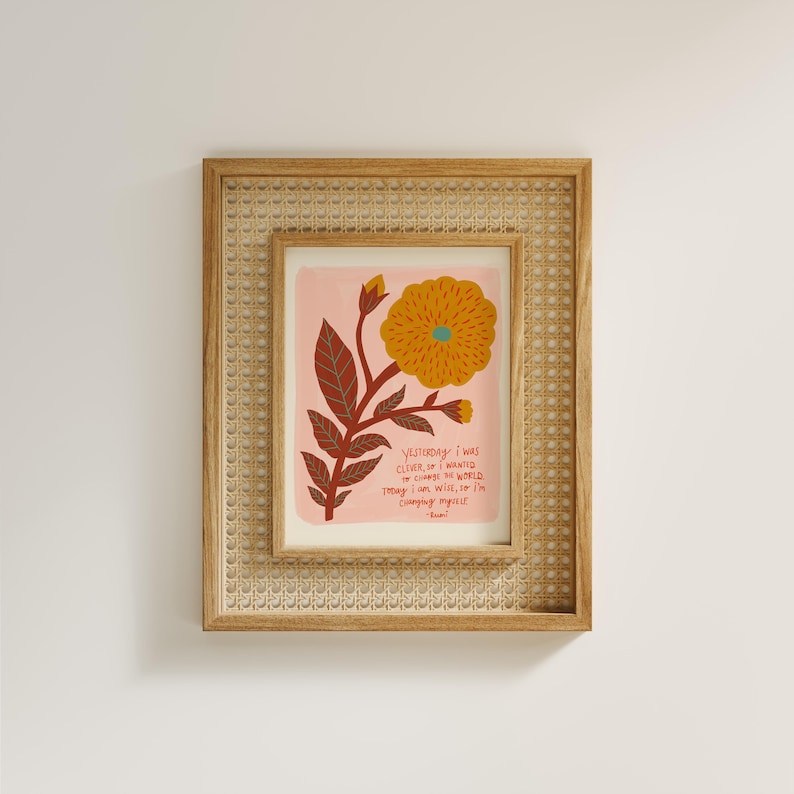 Puede incluir: Una pieza de arte mural enmarcada con fondo rosa claro, una flor grande amarilla, hojas de color rojo oscuro y el texto: Yesterday I was clever, so I wanted to change the world. Today I am wise, so I am changing myself. Rumi. El marco de madera clara tiene una incrustaci&oacute;n de rejilla tejida.