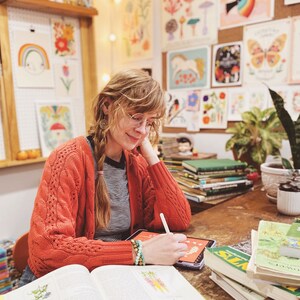 May include: A woman wearing a red cardigan and glasses is drawing on a tablet. She is surrounded by books and artwork featuring nature and plants. The books are about edible plants and wild plants.