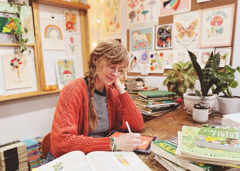 May include: A woman wearing a red cardigan and glasses is drawing on a tablet. She is surrounded by books and artwork featuring nature and plants. The books are about edible plants and wild plants.