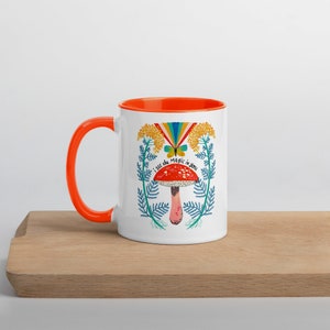 May include: White ceramic mug with an orange handle and interior. The mug features a whimsical design with a red mushroom, butterfly, rainbow, and the text "I see the magic in you". The mug is on a wooden surface.