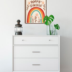 May include: A framed art print with the words "Poppy Sunshine" above a rainbow design. The print is displayed above a white dresser with three drawers. A vintage-style lantern and glass vases with green leaves are on the dresser.