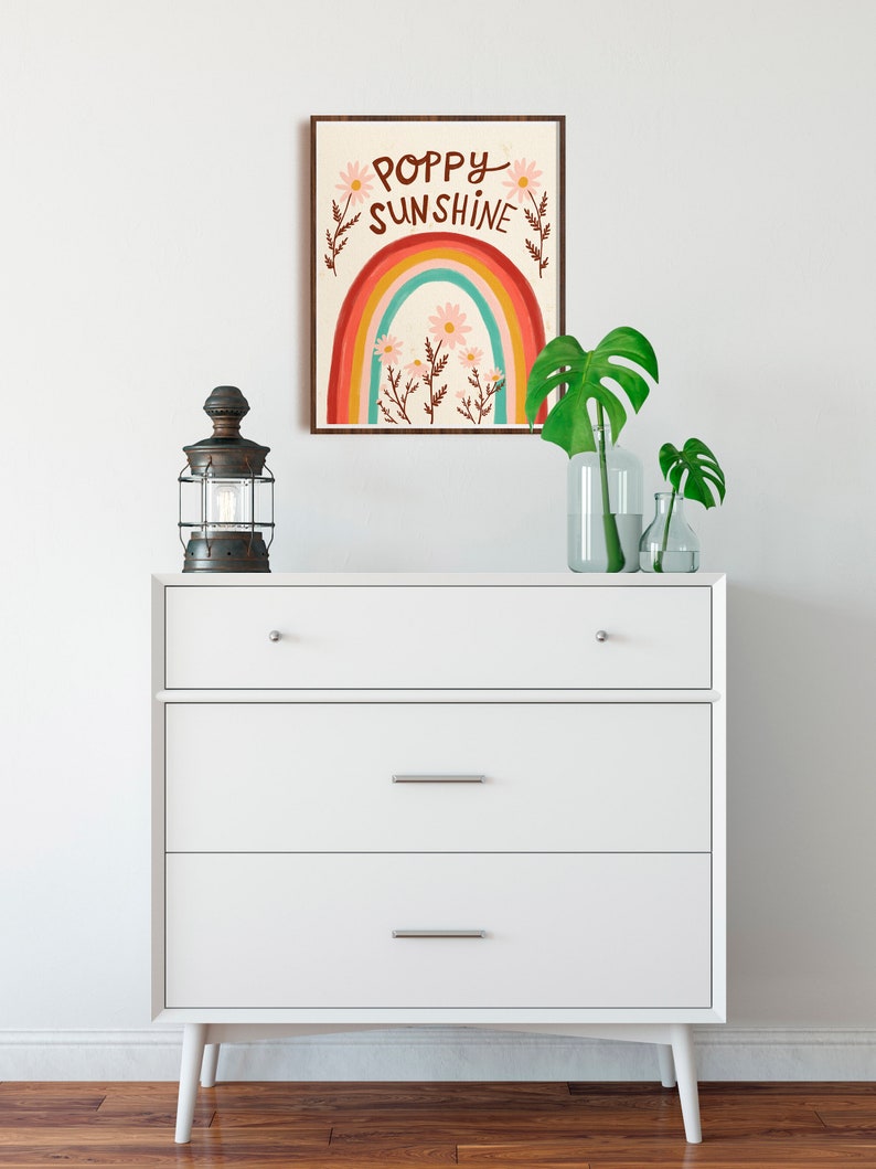 May include: A framed art print with the words "Poppy Sunshine" above a rainbow design. The print is displayed above a white dresser with three drawers. A vintage-style lantern and glass vases with green leaves are on the dresser.